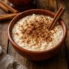 Arroz con leche servido en un tazón de barro artesanal, espolvoreado con canela y decorado con una rama de canela sobre una mesa de madera rústica.