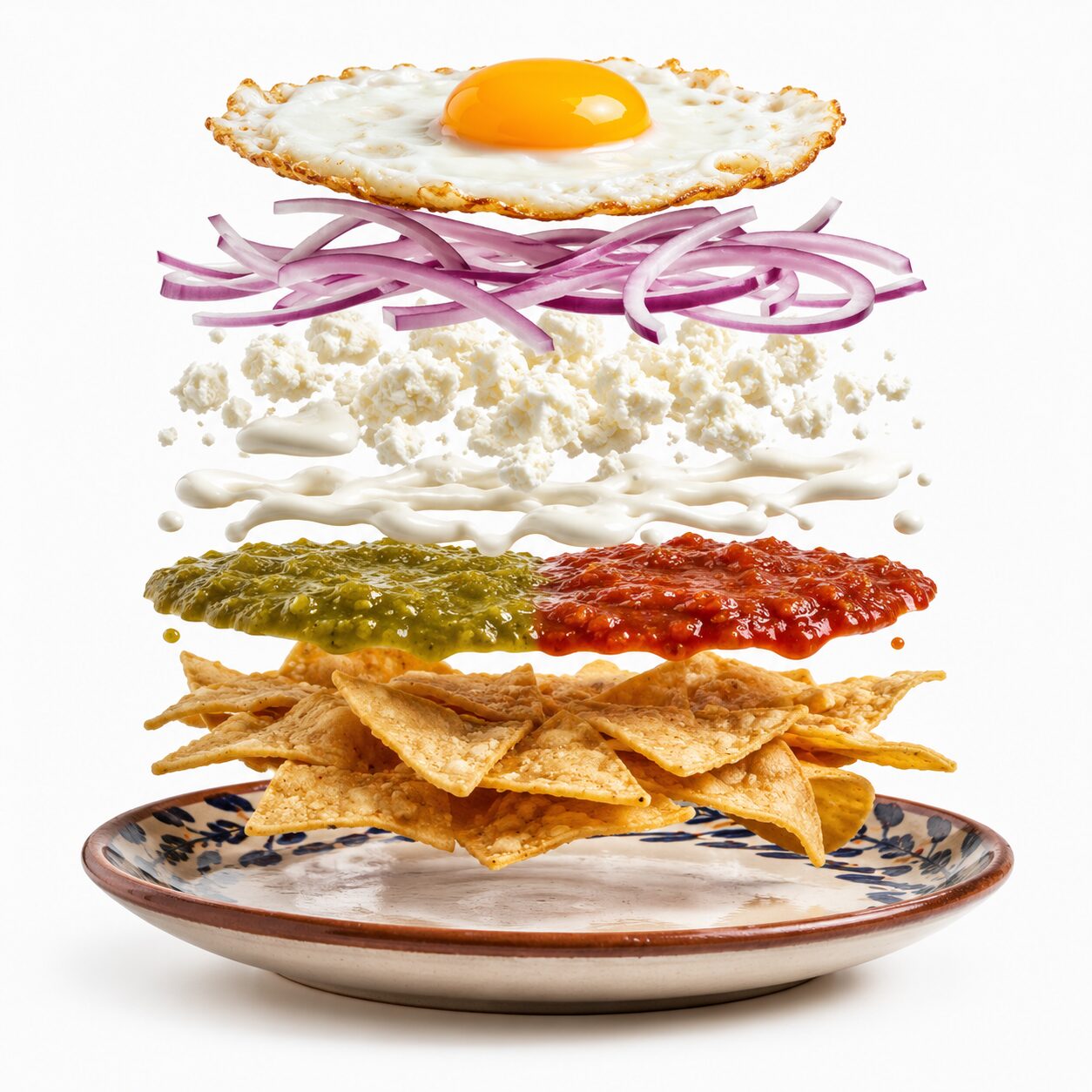 Chilaquiles mexicanos en plato con ingredientes flotando: totopos, salsa verde y roja, crema, queso, cebolla y huevo estrellado sobre fondo blanco.