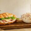 Cuernito de jamón y queso con lechuga, mayonesa y sopa fría de codito servido sobre tabla de madera rústica.