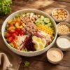 Ensalada fresca en tazón blanco con lechuga, betabel, jícama, zanahoria, tomate cherry, pepino, maíz, durazno, pollo, atún, jamón con queso, huevo cocido y aderezos sobre mesa de madera rústica.