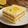 Rebanada de carlota de limón con capas de crema de limón y galleta sobre un plato blanco, decorada con ralladura y una rodaja de limón.